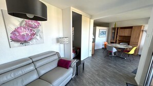 Uma sala de estar moderna com um sofá cinzento, uma área de jantar com mesa redonda e cadeiras, e uma cozinha ao fundo.