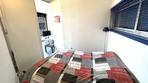 Um quarto com cama, escrivaninha, cadeira, televisão e janela com persianas.