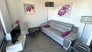 Uma sala de estar com um sofá cinzento, duas almofadas cor-de-rosa, uma mesa pequena, um abajur e duas pinturas penduradas na parede.