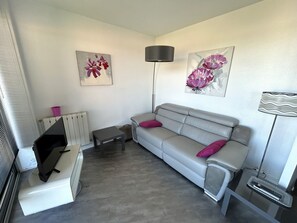 A living room with a grey sofa, two pink cushions, a small table, a floor lamp, and two wall-mounted paintings. - Apartment with AC, panoramic view - 4 guests (Argelès-sur-Mer)