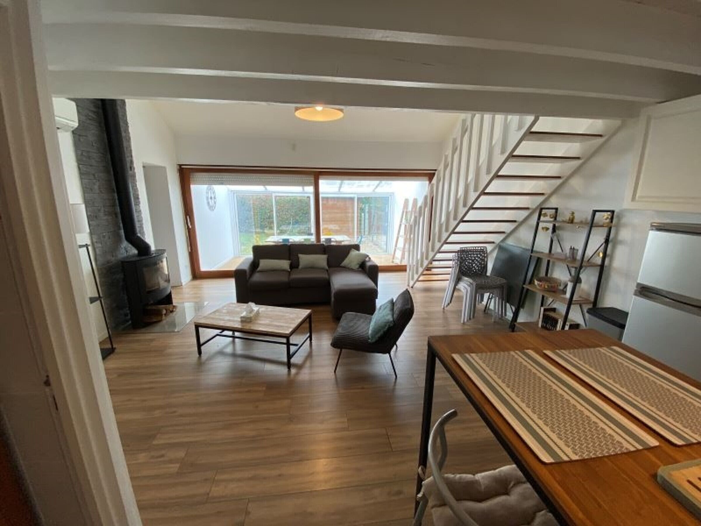 A spacious living area with a sofa, coffee table, and a dining table with placemats. A fireplace is visible on the left, and a staircase leads to an upper level.
