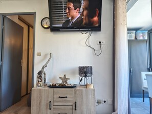 A modern living room with a television mounted on the wall, a clock, a decorative star, a lamp, and a wooden cabinet. - Air-conditioned apartment, parking - 4 guests near the port (Gruissan)