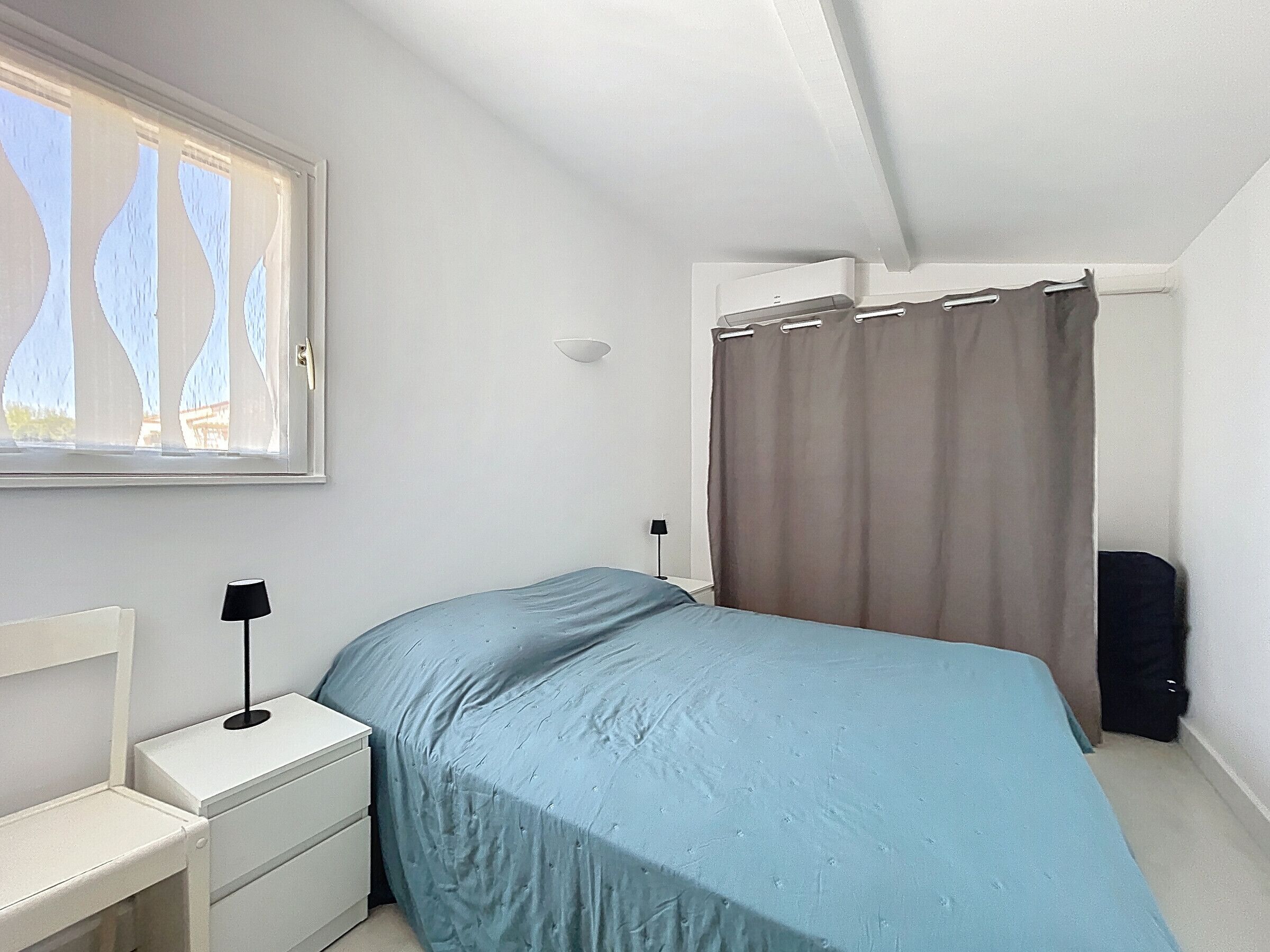 Un dormitorio con cama, mesita de noche, lámpara, ventana con cortinas y una puerta.