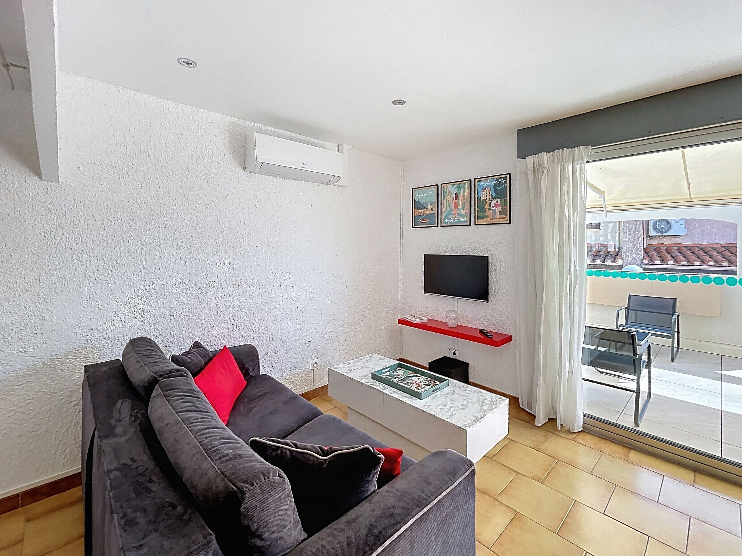 Sala de estar com sofá cinza, televisão de tela plana, mesa de centro pequena e varanda com vista.