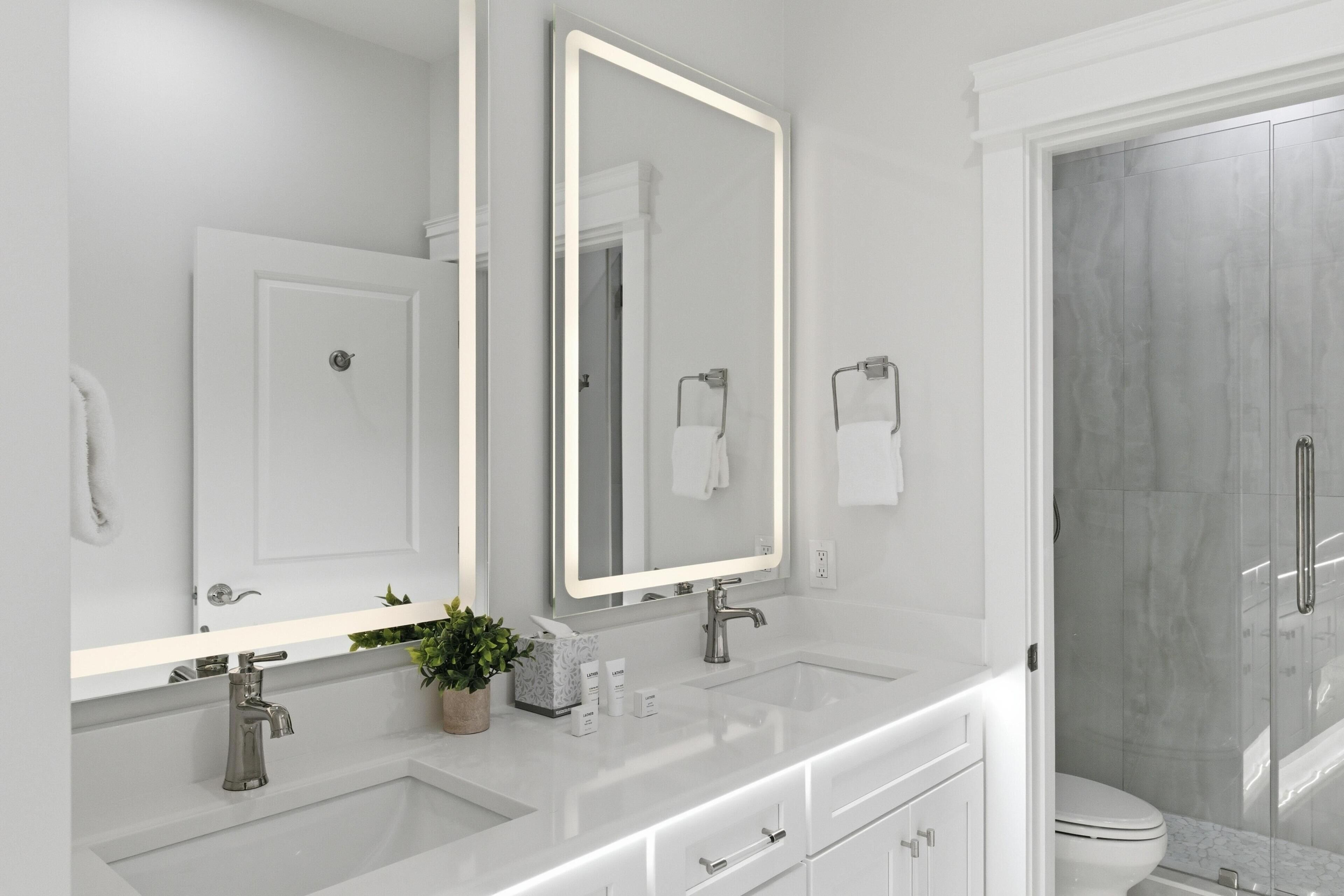 A modern bathroom with a large mirror, two sinks, a shower, and a toilet.