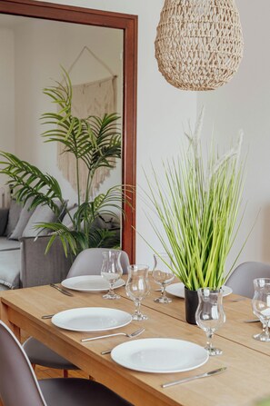Une table à manger dressée avec des assiettes, des verres et des couverts, un luminaire tressé suspendu au-dessus, et une plante en pot aux longues feuilles vertes.