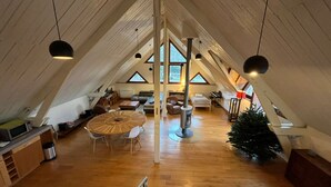 Un ático espacioso con techo alto, vigas de madera y chimenea central. Hay una mesa de comedor con sillas, un sofá y un árbol de Navidad.