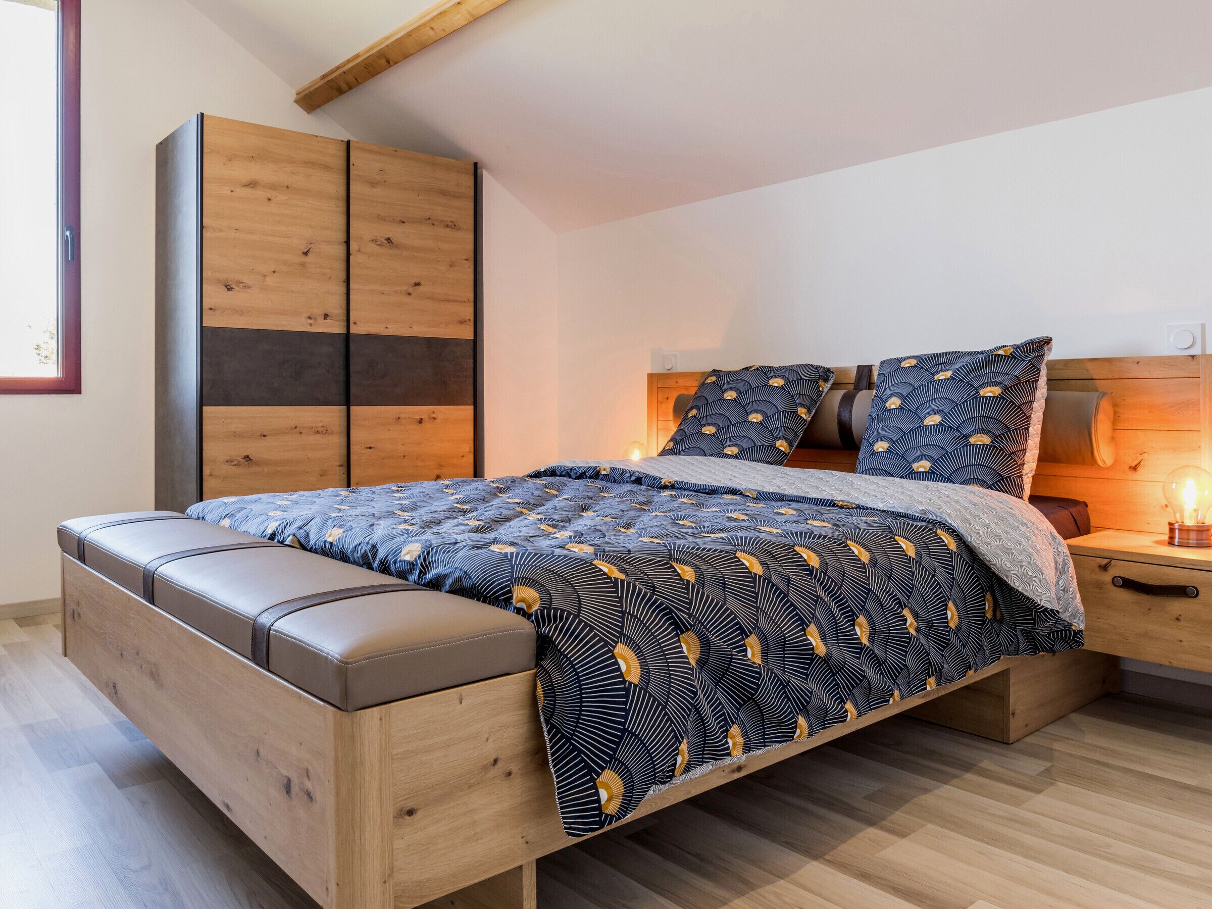 A bedroom with a bed, wooden wardrobe, bedside table, and a nightstand with a lamp.