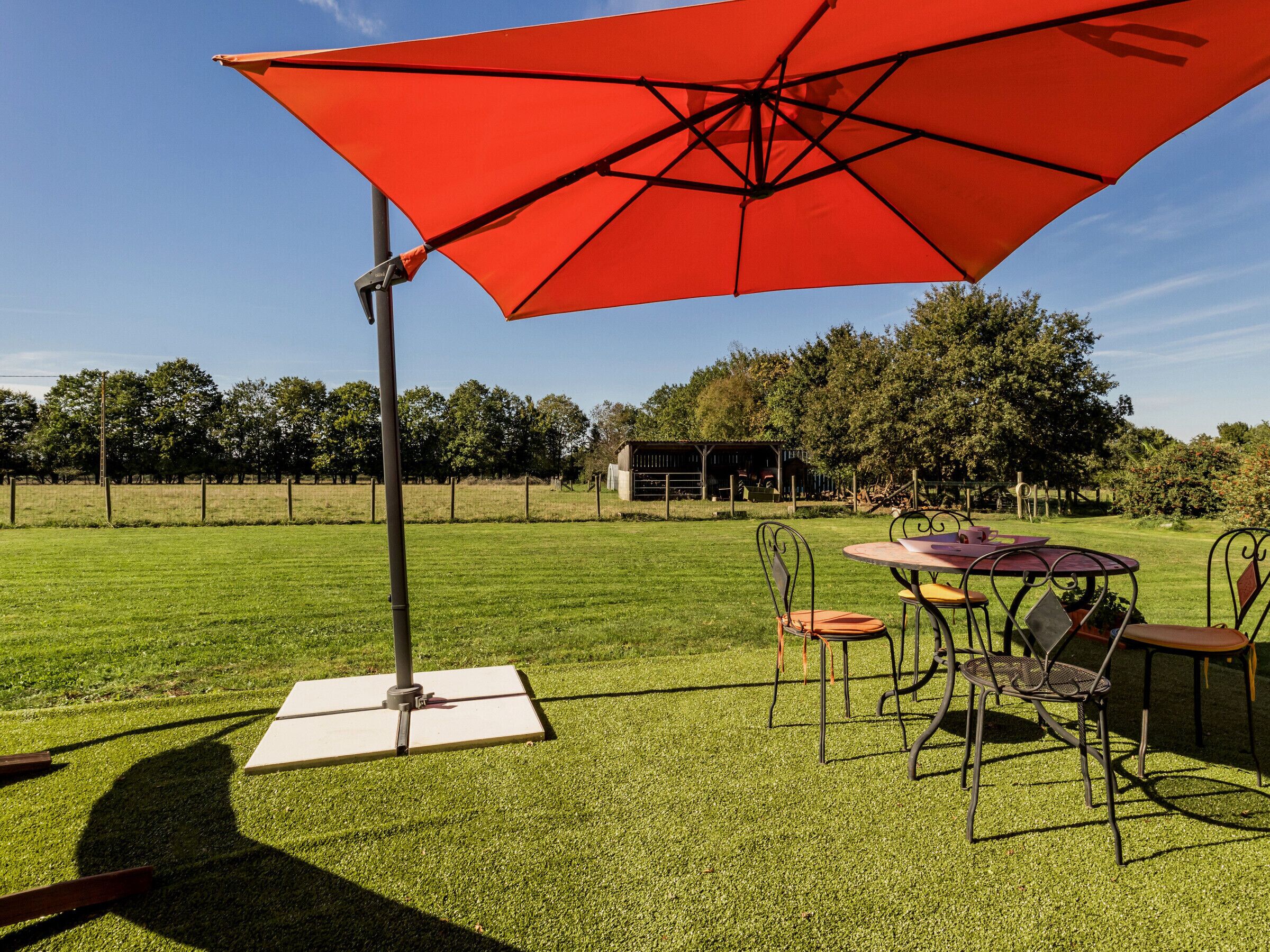 An outdoor patio with a table and chairs under a red umbrella, a white tablecloth, and a metal fence in the background.