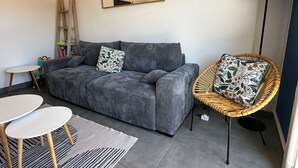 Een grijze bank met gekleurde kussens, een rieten stoel, een klein rond tafeltje, een vloerlamp en een spiegel aan de muur.