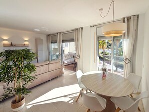 Een moderne woonkamer met een bank, een eettafel met stoelen, een grote potplant en uitzicht op het buiten.