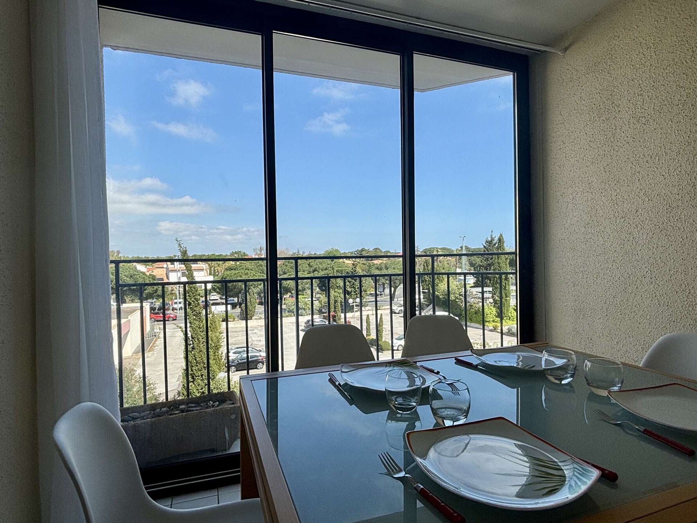 Ein Essbereich mit einem Tisch für vier Personen, große Fenster mit Blick auf einen Parkplatz und Gebäude, sowie ein Balkon mit Geländer.