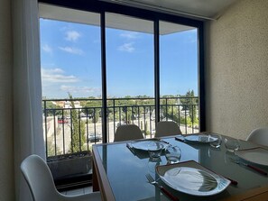 A dining area with a table set for four, large windows with a view of a parking lot and buildings, and a balcony with a railing. - Apartment near sea, elevator - 4 guests (Argelès-sur-Mer)