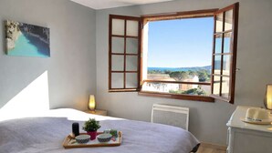 A bedroom with a bed, a window with open shutters, a bedside table with a lamp, and a tray with bowls and a plant. - Quiet apartment, sea view - 4 guests (Saint-Aygulf)