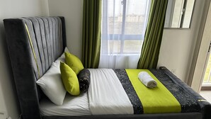 Uma cama com cabeceira cinzenta, roupa de cama branca e verde, e uma toalha enrolada.
