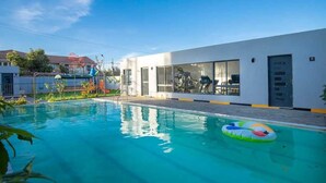 Uma piscina com um flutuador colorido, um edifício branco com grandes janelas e equipamentos de ginástica visíveis através delas.