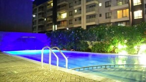 Uma piscina com escada de metal, rodeada por um pátio de tijolos. Ao fundo, um edifício com vários andares com varandas e janelas.