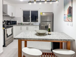 Une cuisine moderne dotée d’un plan de travail en granit, d’appareils électroménagers en acier inoxydable et d’une table en bois accompagnée de deux tabourets blancs.