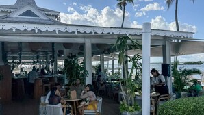 Een openluchtrestaurant met een overdekt terras, palmbomen en uitzicht op het water.