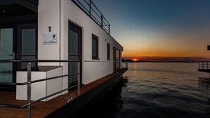 A modern building with a sign reading 'Kranich West' by a calm body of water at sunset. - KranichNest Schwimmhaus Uferhaus am Bodden – mit Terrasse & Seeblick für 4 Pers. (Ribnitz-Damgarten)
