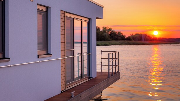 A modern building with large windows, a balcony, and a view of a sunset over water. - KranichNest Schwimmhaus Uferhaus am Bodden – mit Terrasse & Seeblick für 4 Pers. (Ribnitz-Damgarten)