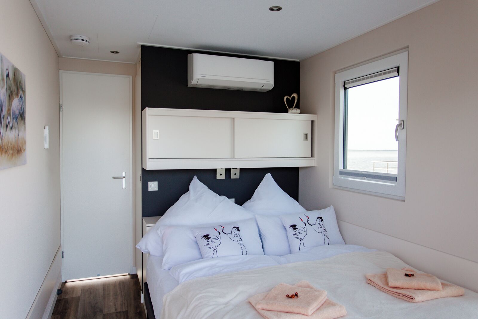 Een slaapkamer met een bed, witte kussens, een raam, een airconditioning en een deur.