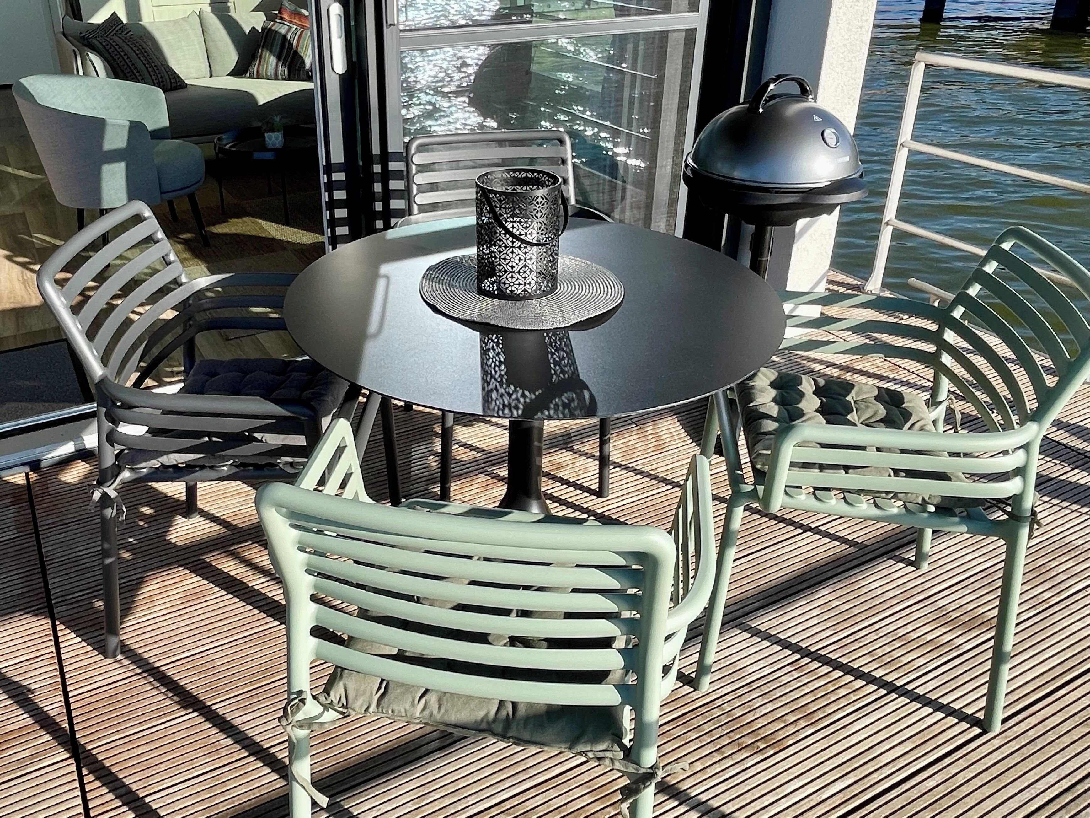 Ein runder Tisch mit einem gemusterten Mittelteil, umgeben von grünen Metallstühlen auf einer Holzterrasse mit Blick auf das Wasser.