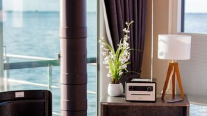 A modern living room with a wood-burning stove, a view of the sea, a potted plant, a radio, and a lamp on a wooden cabinet. - KranichNest Schwimmhaus Uferhaus am Bodden – mit Terrasse & Seeblick für 4 Pers. (Ribnitz-Damgarten)