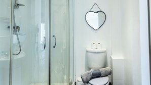 Kylpyhuoneessa on lasiseinäinen suihkukaappi, wc-istuin, jonka päällä on pyyhe, ja sydämenmuotoinen peili seinällä.