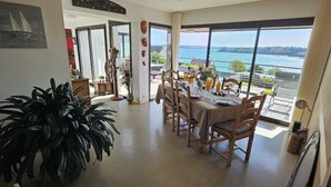 Un coin repas avec une table dressée pour quatre personnes, une grande plante en pot et une vue sur la mer grâce à de grandes fenêtres.