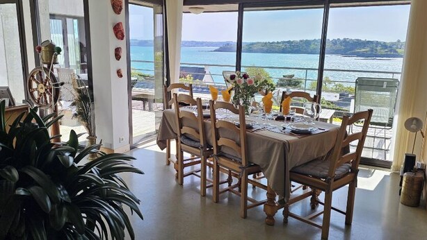 Un coin repas avec une table dressée pour quatre personnes, de grandes fenêtres offrant une vue sur la mer et une plante en pot au premier plan.