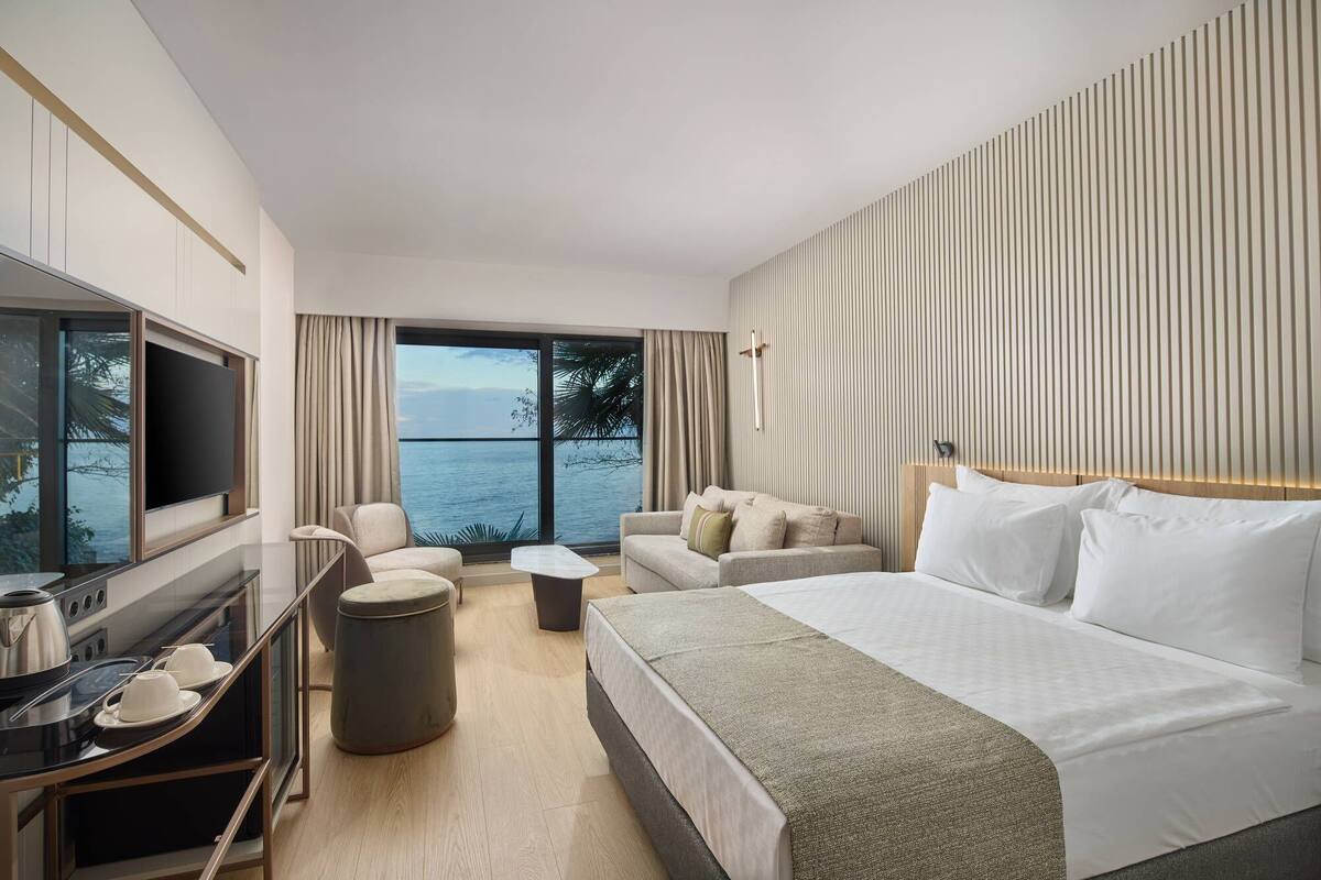 A hotel room with a bed, a sofa, a TV, a desk with a kettle, and a view of the sea.