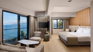 A modern hotel room with a large bed, a seating area with a small table, a TV, and a view of the sea.