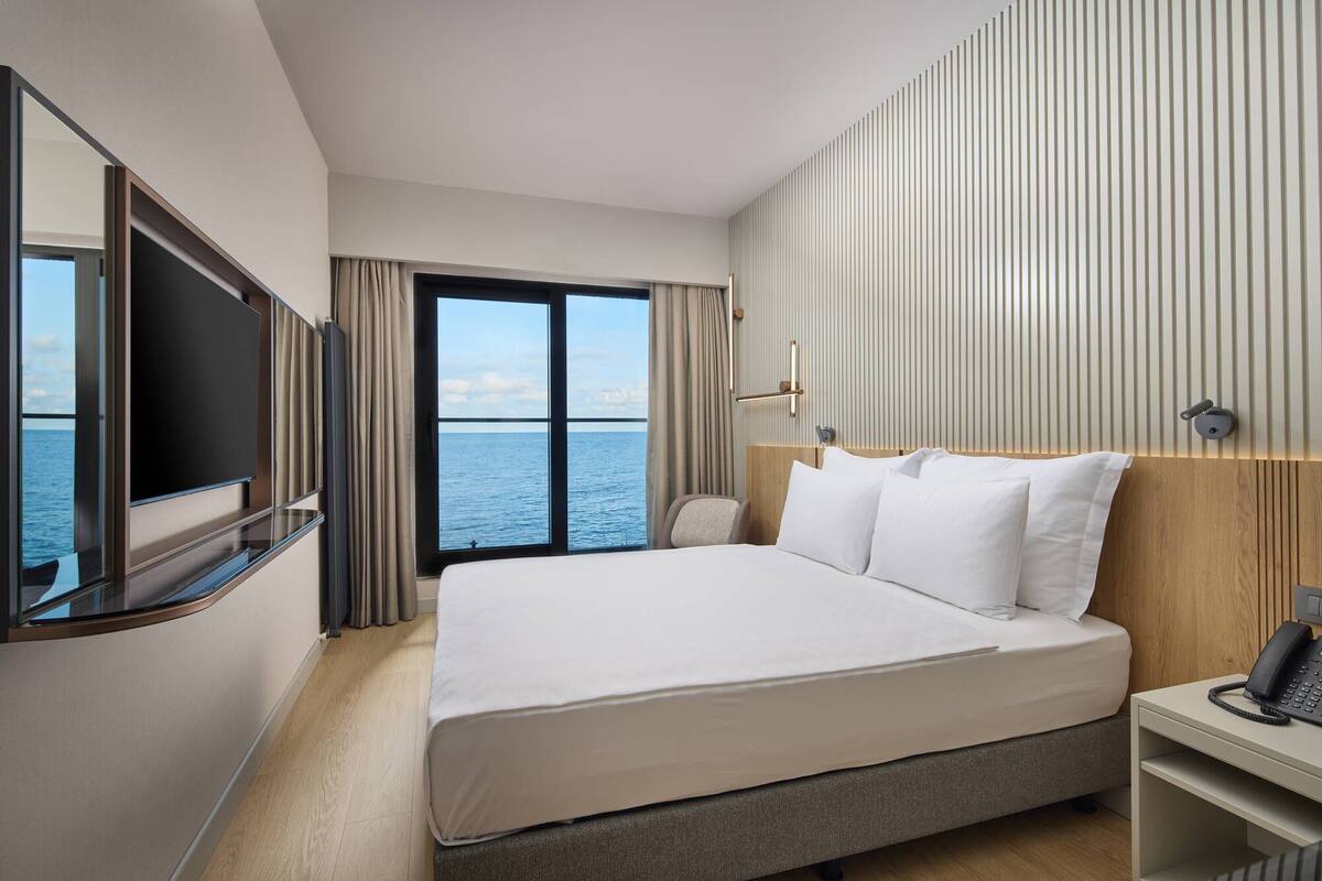 A hotel room with a large bed, a TV, a bedside table with a phone, and a view of the sea.