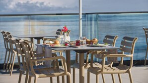 A dining table with chairs, a pitcher, glasses, and plates of food, set against a view of the sea.