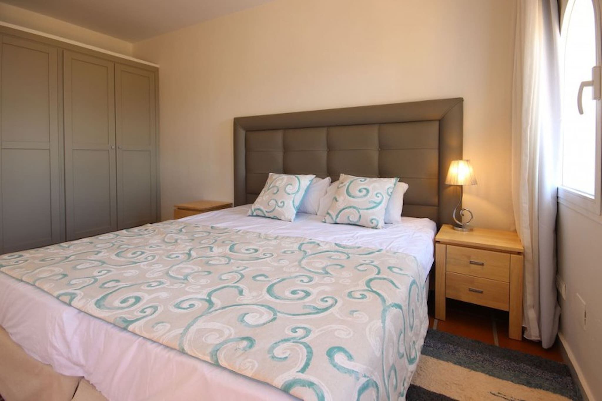 A bedroom with a bed, a nightstand with a lamp, a wardrobe, and a window with curtains.