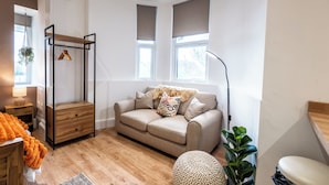 A room with a wooden floor, a beige sofa, a small wooden nightstand, a hanging plant, and a window with blinds. - Glan Neigr by Guestz (Rhosneigr)