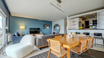 A modern living room with a dining area, a sofa, a TV, and a kitchen in the background.