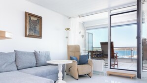 Ein modernes Wohnzimmer mit Sofa, Sesseln und einem Tisch, einem Gemälde an der Wand und Blick auf das Meer durch große Glasfenster.