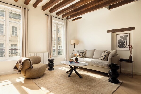 A living room with a sofa, armchair, coffee table, and a view of buildings through large windows. - Marais Gem 2BR 6P Paris Stay (Paris)