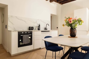 A modern kitchen with white cabinetry, a black oven, and a dining area with a white table and blue chairs. - Marais Gem 2BR 6P Paris Stay (Paris)