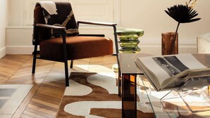 Una habitación con un ventanal grande, una silla, una mesa de vidrio y un florero con flores.
