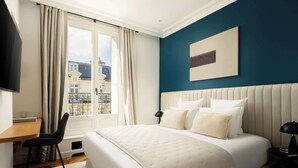 Habitación de hotel con cama, escritorio, silla, ventana con cortinas, televisor montado en la pared y un cuadro.