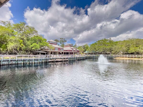A serene lake with a fountain, surrounded by lush trees and a bridge leading to a building with a red roof. - Oceanfront Turtle Dunes 5th Floor RENOVATED - Private Balcony - Beach & Pool! (Fernandina Beach)