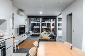 A modern living space with a kitchen, dining area, and seating area, featuring a large window, built-in appliances, and a flat-screen TV. - Upper West Side Spencer Heights 2BR 1Car (Melbourne)