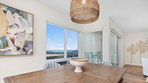Uma sala de jantar com mesa de madeira, cadeiras listradas, luminária trançada, um quadro abstrato grande e vista para o oceano através de janelas amplas.
