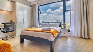 A modern hotel room with a large bed, a desk, a chair, a nightstand, and a view of the cityscape through a large window. - Cozy studio apartment in lovely Nairobi with amazing city views (Nairobi)