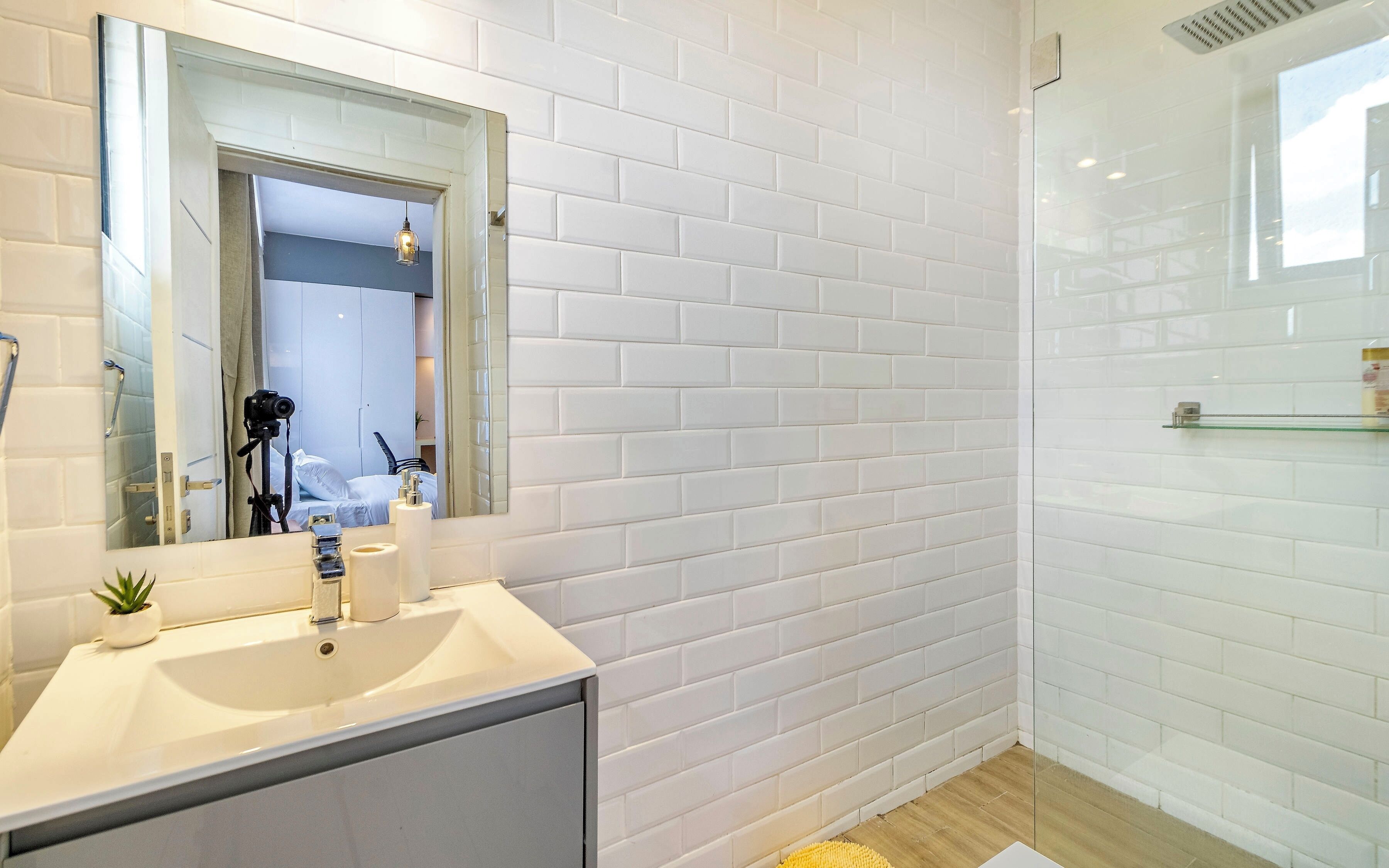 A bathroom with white tiled walls, a mirror, a sink, and a shower area.
