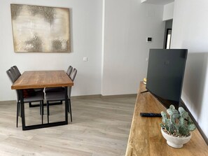 A modern living room with a wooden dining table, grey chairs, a flat-screen TV, and a cactus plant on a wooden side table. - Estepona Center, Pool and Beach (Estepona)