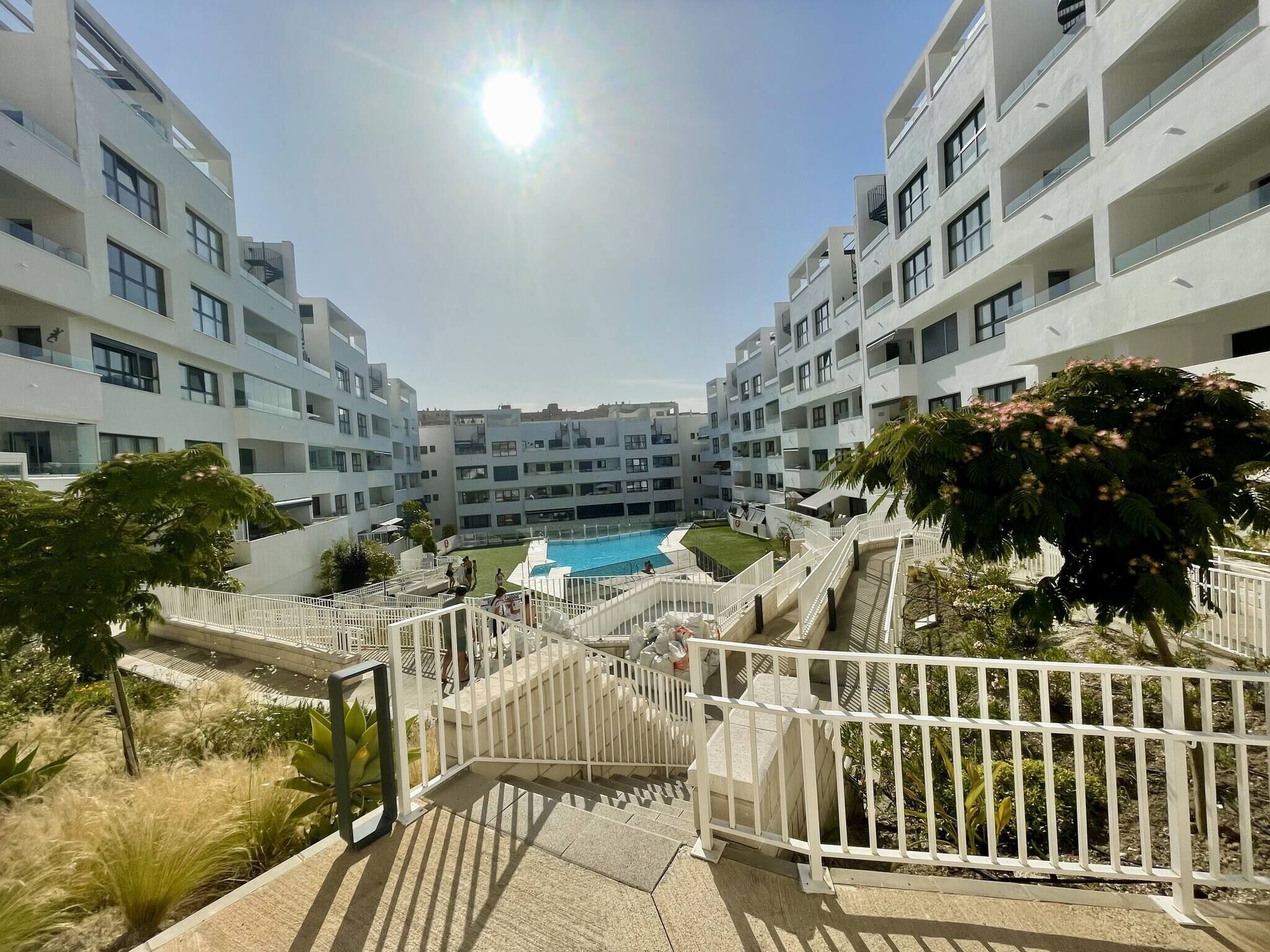 A modern apartment complex with a central swimming pool, white balconies, and a clear sky.